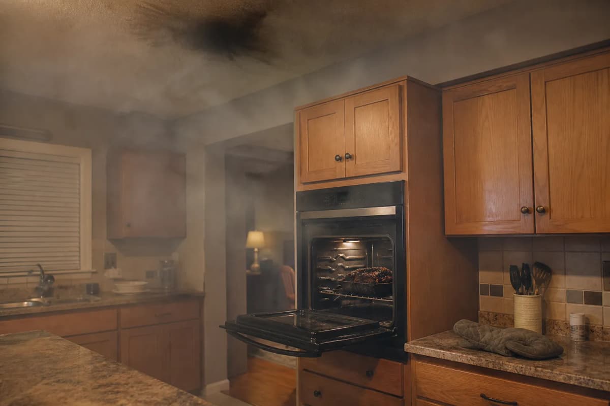 A smoke-filled kitchen with an open oven and a burned pan inside.